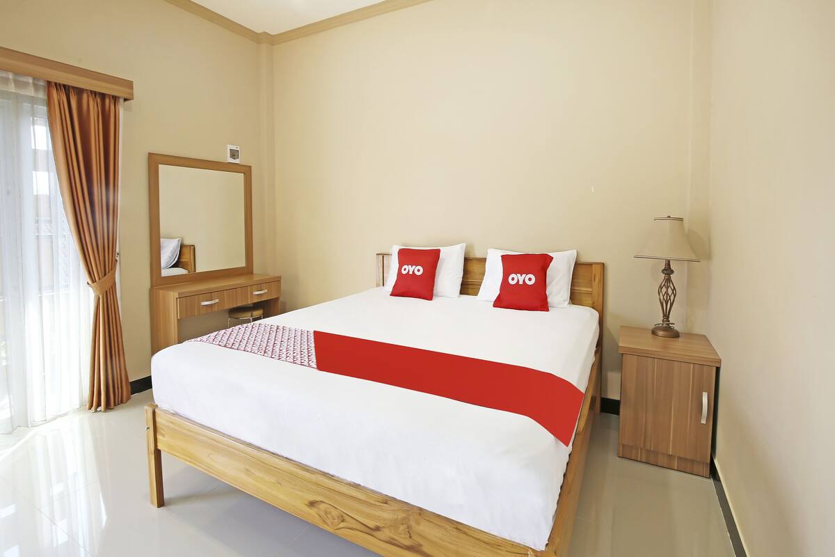 A hotel room with a bed, a wooden nightstand, a mirror, and a lamp.