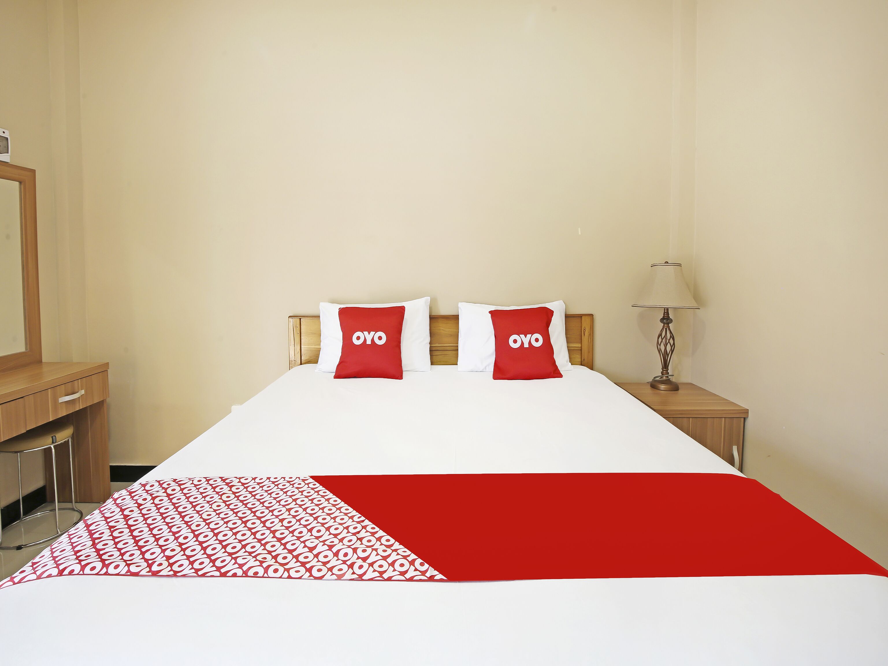 A hotel room with a bed, two pillows with the word "OYO" on them, a bedside table with a lamp, and a wooden nightstand.