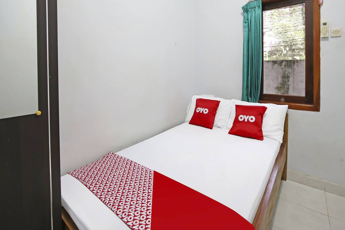 A bed with white bedding and red pillows, a window with teal curtains, and a visible wall-mounted light switch.