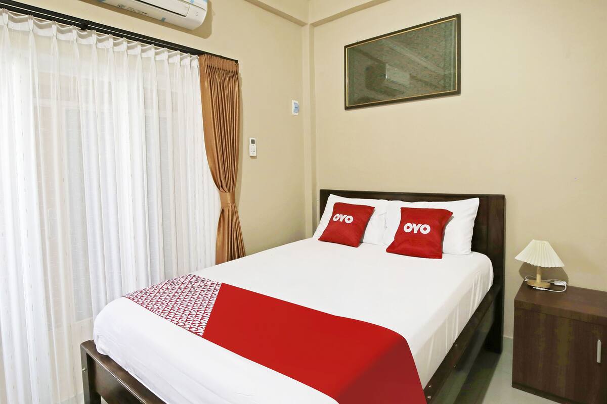 A neatly made bed with a white and red bedspread, two red pillows with the word "OYO" on them, a wooden nightstand with a lamp, and a painting on the wall.