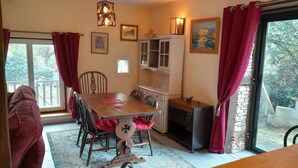 A dining area with a wooden table, chairs, a red sofa, a small table with a radio, a fireplace, and a view of the outdoors through a glass door. - Charming detached country cottage (Salagnac)
