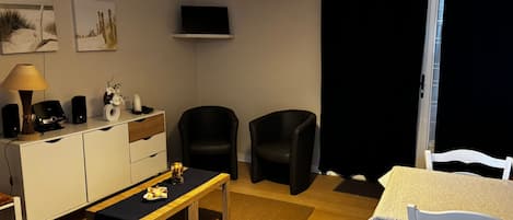 A modern living room with a flat-screen TV, a dining table, two chairs, a sofa, a coffee table, and a lamp.
