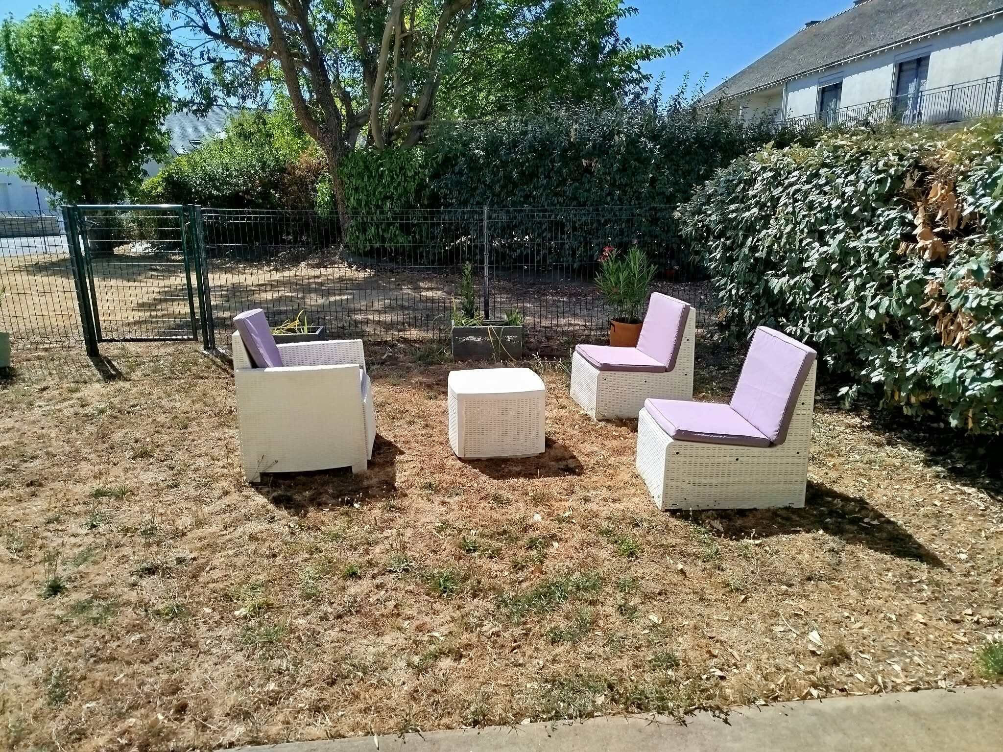 An outdoor seating area with three chairs and a small table, surrounded by dry grass and a metal fence.