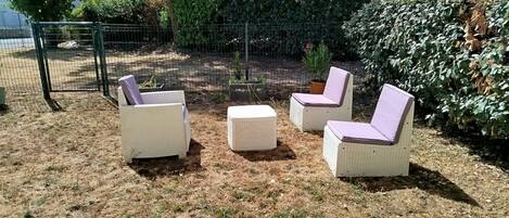 An outdoor seating area with three chairs and a small table, surrounded by dry grass and a metal fence.