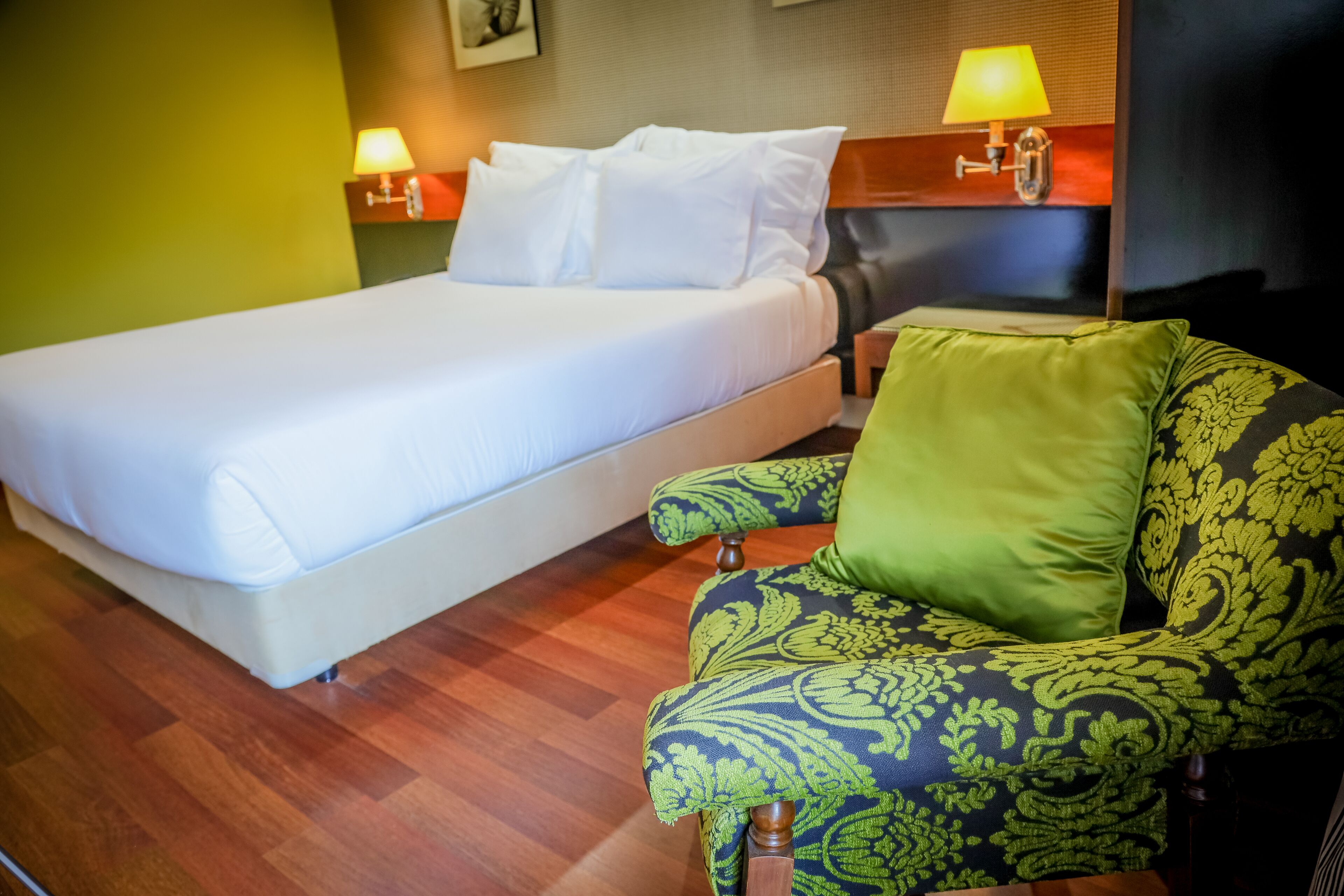 A hotel room with a bed, a green and black patterned armchair, and two bedside lamps.