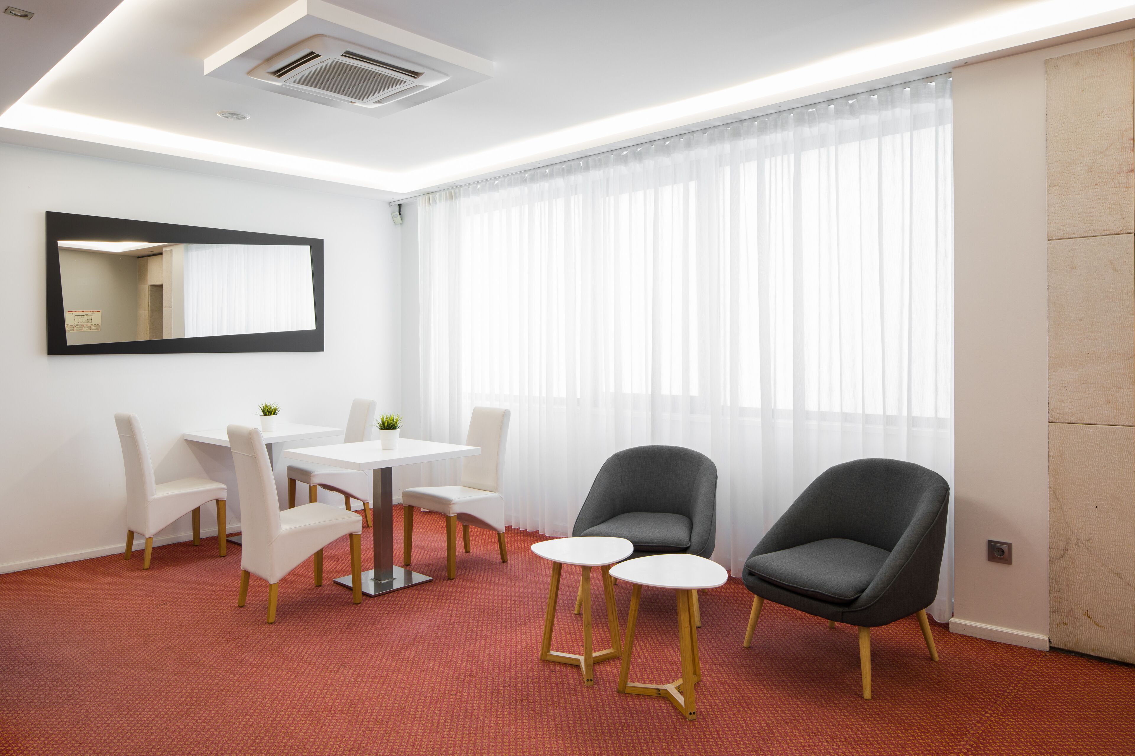 Habitación de hotel con comedor, un espejo grande, una ventana con cortinas traslúcidas y piso cubierto de alfombra roja.