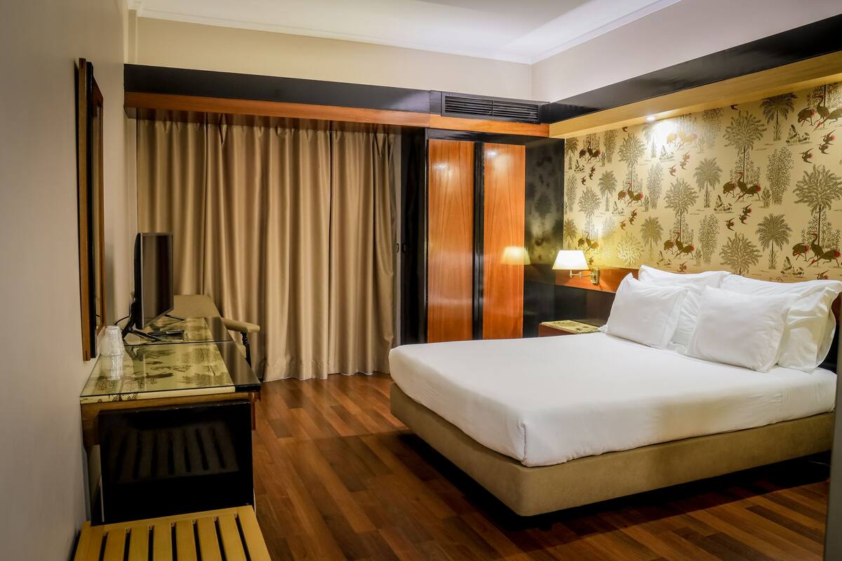 A hotel room with a bed, a desk, a chair, a wardrobe, and a wall with a floral pattern.