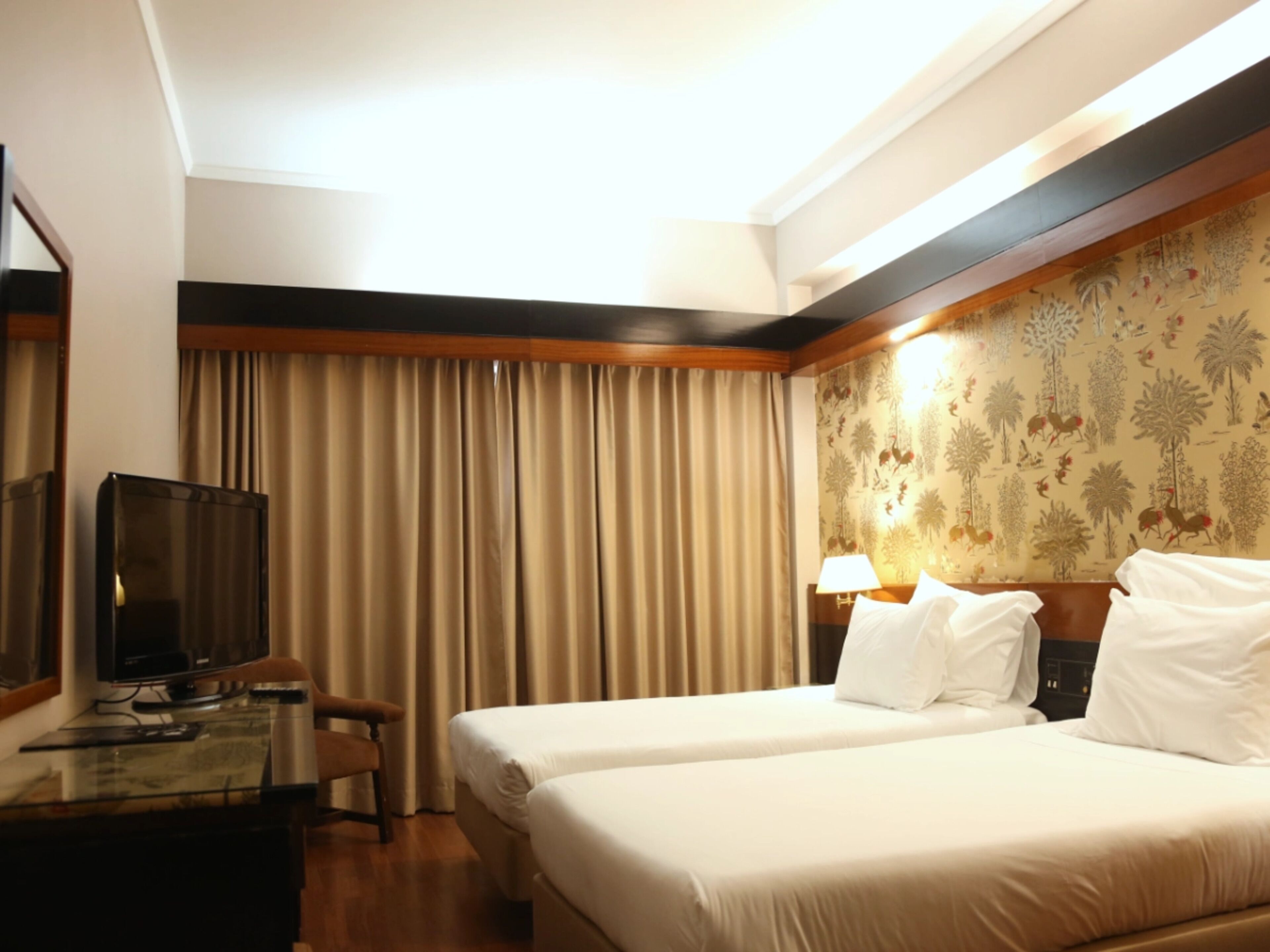 A hotel room with two beds, a desk with a TV, and a large window with curtains.