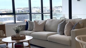 Una sala de estar moderna con un sofá color beige, una mesita redonda de madera y vistas a una zona residencial a través de amplios ventanales.