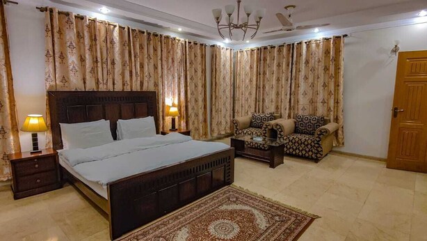 A bedroom with a bed, two bedside tables with lamps, a patterned sofa, a patterned rug, and a wooden door. - Dream Home (Islamabad)