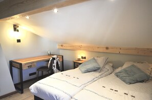 A bedroom with a bed, a desk, and a chair. There are wall-mounted lights and a wooden ceiling beam.