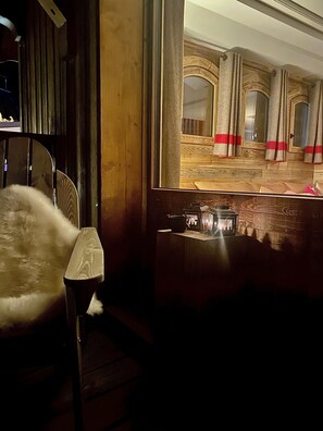 A dimly lit room with a large mirror reflecting an interior space with wooden flooring and red curtains. A chair with a white cushion is positioned in front of the mirror.