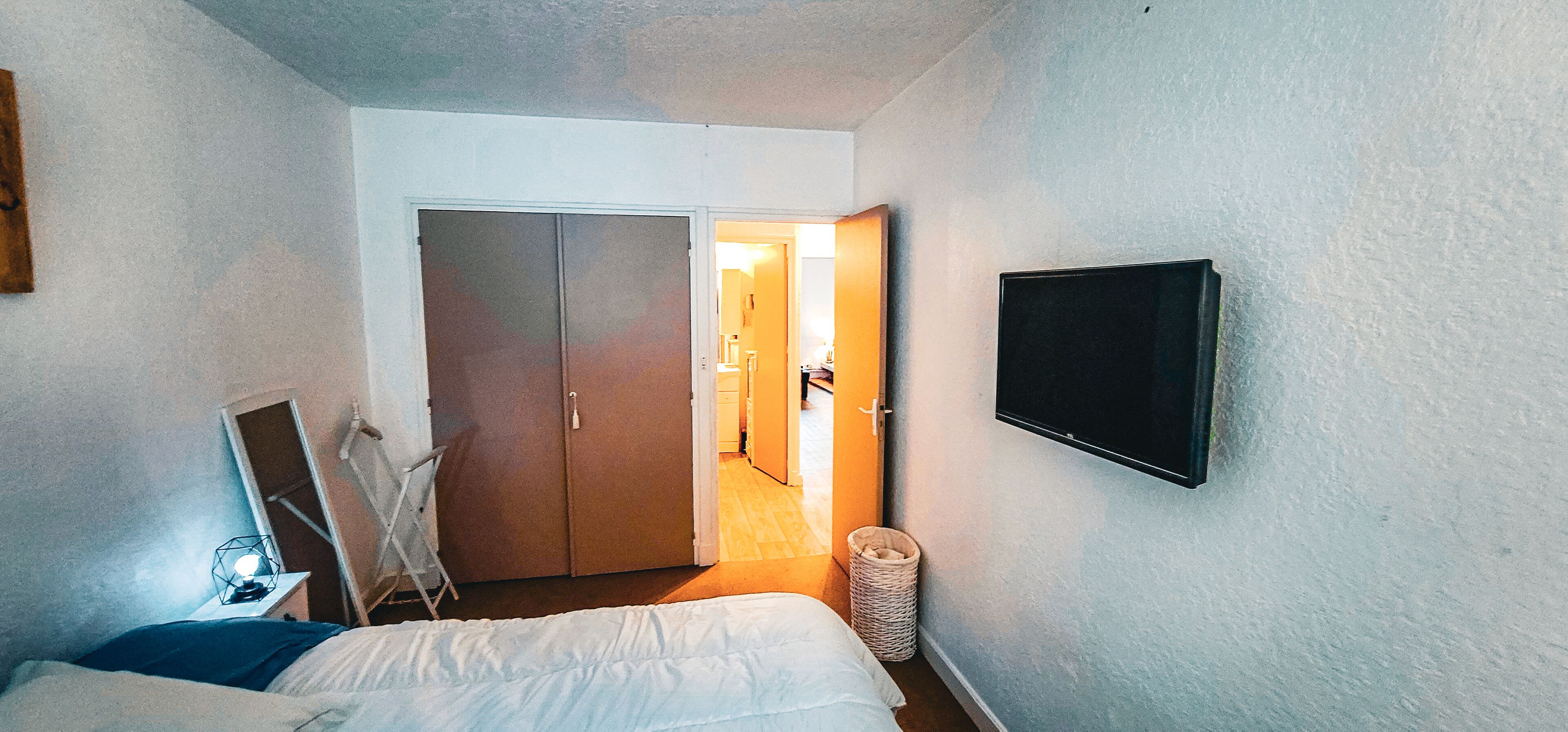 Ein Schlafzimmer mit einem Bett, einem an der Wand montierten Fernseher, einem Kleiderschrank und einem kleinen Tisch mit einer Lampe.
