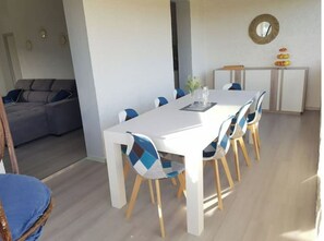 Uma área de refeições com mesa branca e cadeiras azuis, um sofá cinzento e um pequeno armário com objetos decorativos.