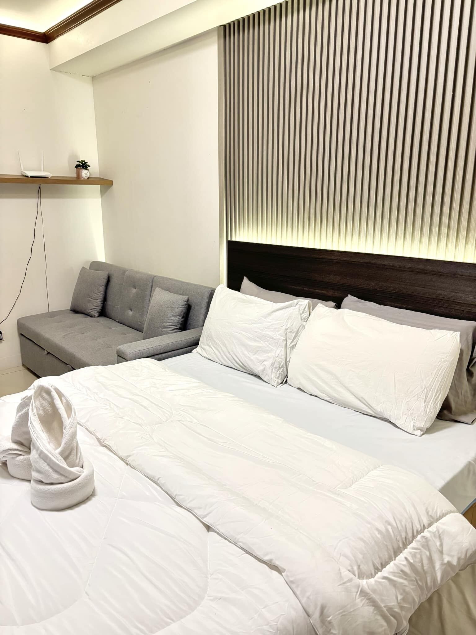 A modern hotel room with a large bed, a grey sofa, and a wooden shelf with a plant and a lamp.
