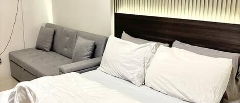 A modern hotel room with a large bed, a grey sofa, and a wooden shelf with a plant and a lamp.