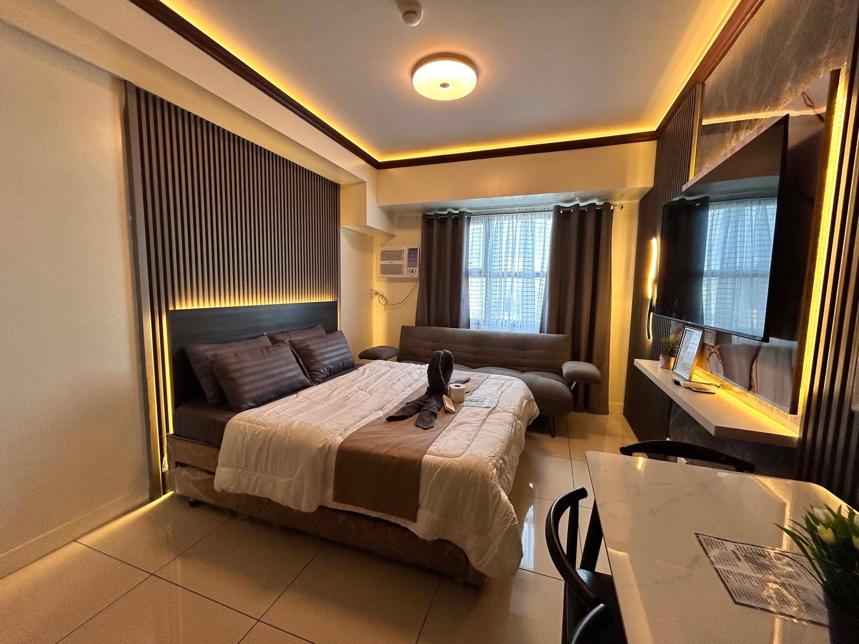 A modern hotel room with a large bed, a desk with a computer, a TV, and a window with curtains.