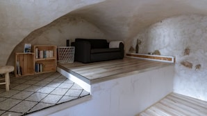 A small, cozy attic room with a sloped ceiling, a black sofa, a wooden bookshelf, and a tiled floor. - Lou Roucas : Pleasant village house with view (Fontaine-de-Vaucluse)