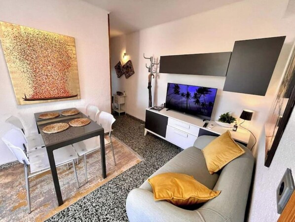 Una sala moderna con televisor de pantalla plana, una mesa de comedor con sillas, un sofá con cojines y un elemento decorativo en la pared.
