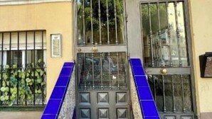 Ein Gebäudeeingang mit einem Metallgitter, der Zahl '2' über der Tür und blautonierten Stufen, die zum Eingang führen.