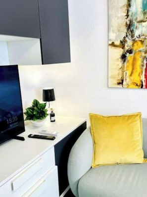 Una habitación moderna con un escritorio blanco, un monitor negro, una planta pequeña, una lámpara y un cojín amarillo sobre un sofá gris.