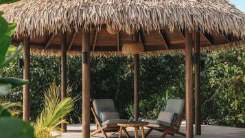Un área de estar al aire libre con techo, dos sillones de relax y una mesita baja, rodeada de abundante vegetación.