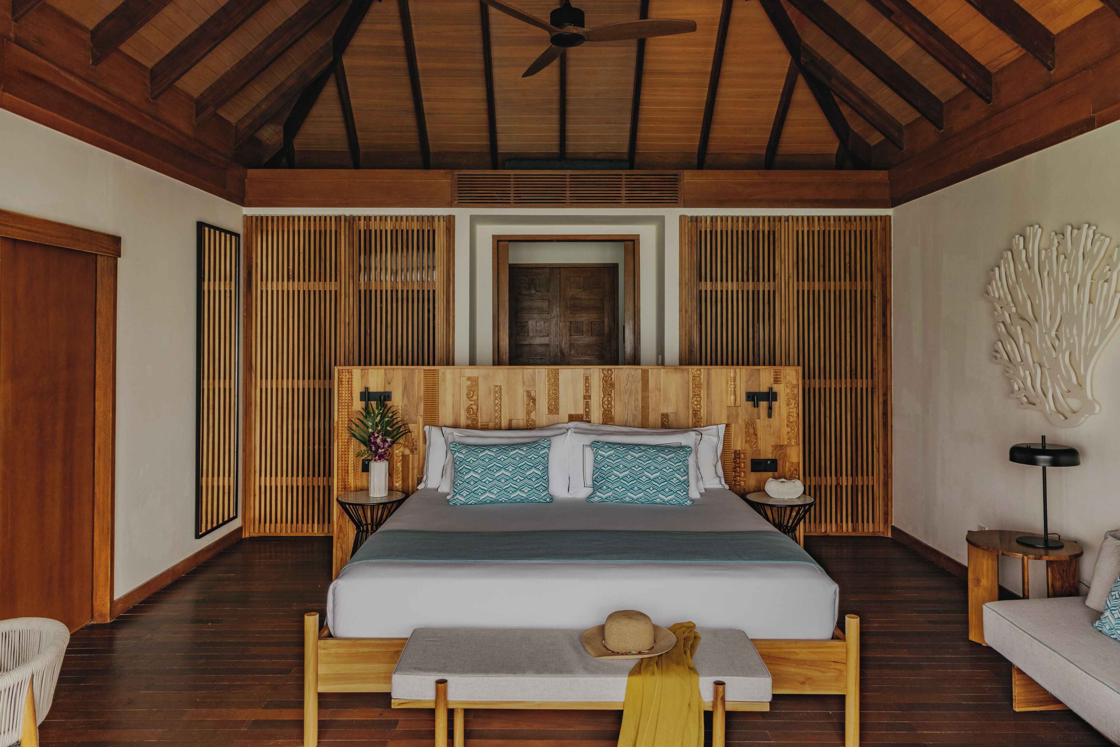 A bedroom with a wooden ceiling, a bed with a grey bedspread, two pillows, a bedside table with a lamp, and a wall-mounted artwork.