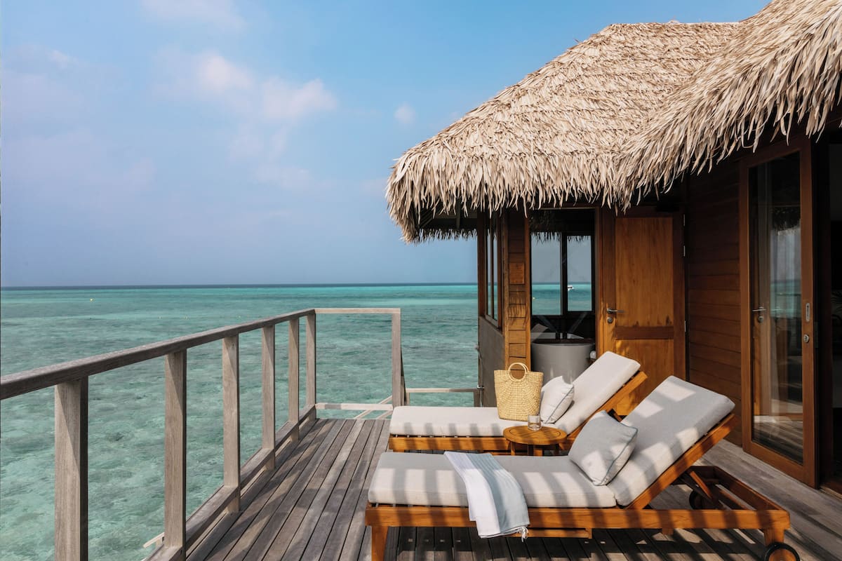 A wooden deck with a thatched roof, a lounge chair, a small table, and a view of the ocean.