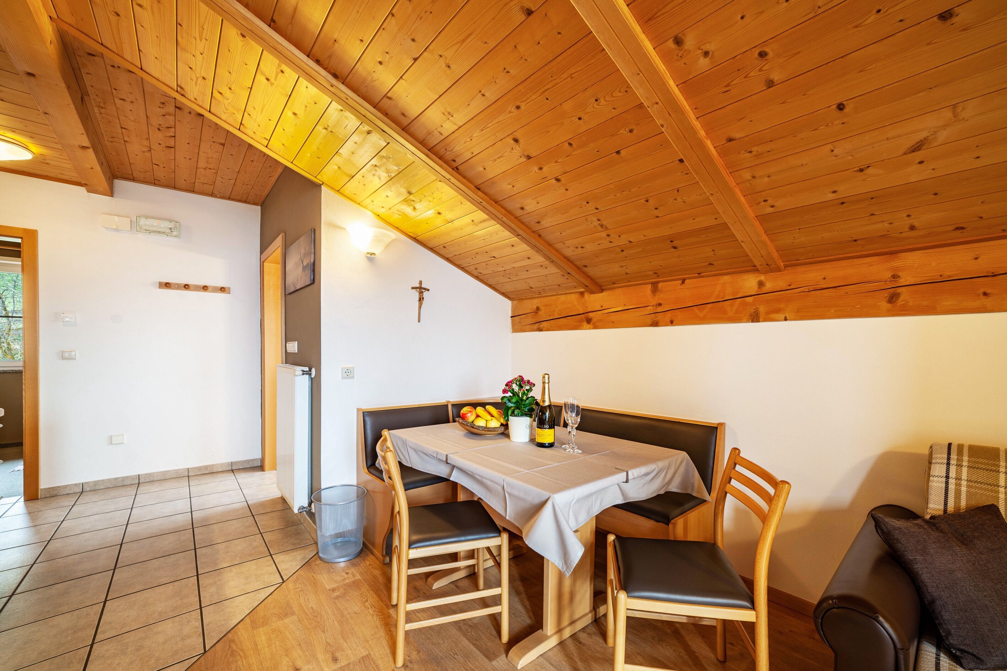 A dining area with a wooden ceiling, a table set with a bottle, glasses, and a bowl of fruit.