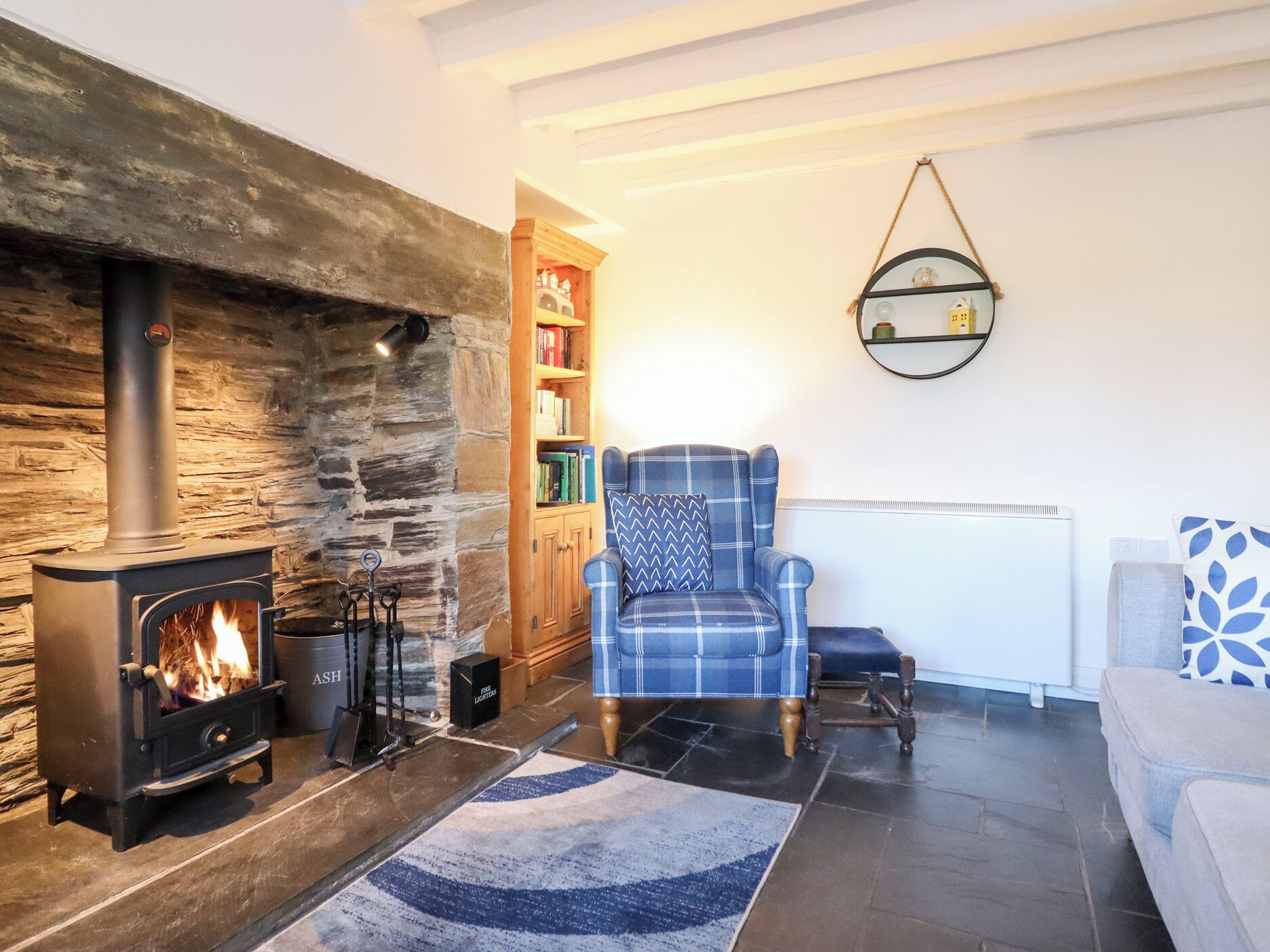 A cozy living room with a stone fireplace, a blue plaid armchair, a bookshelf, and a white radiator.