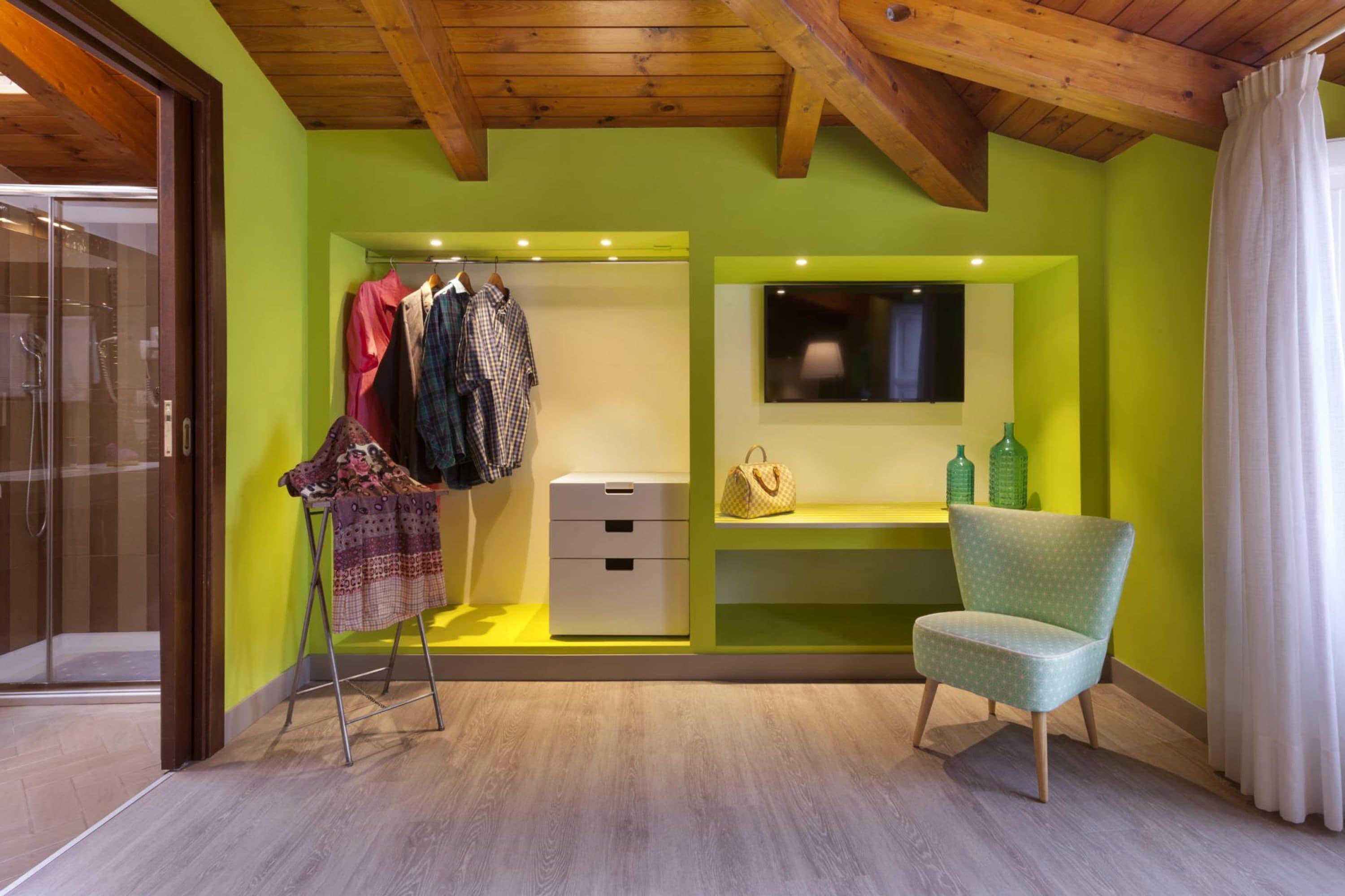 Una habitación con techo de madera, pared verde, suelo de madera, una silla, un perchero, un mueble blanco pequeño y un espejo.