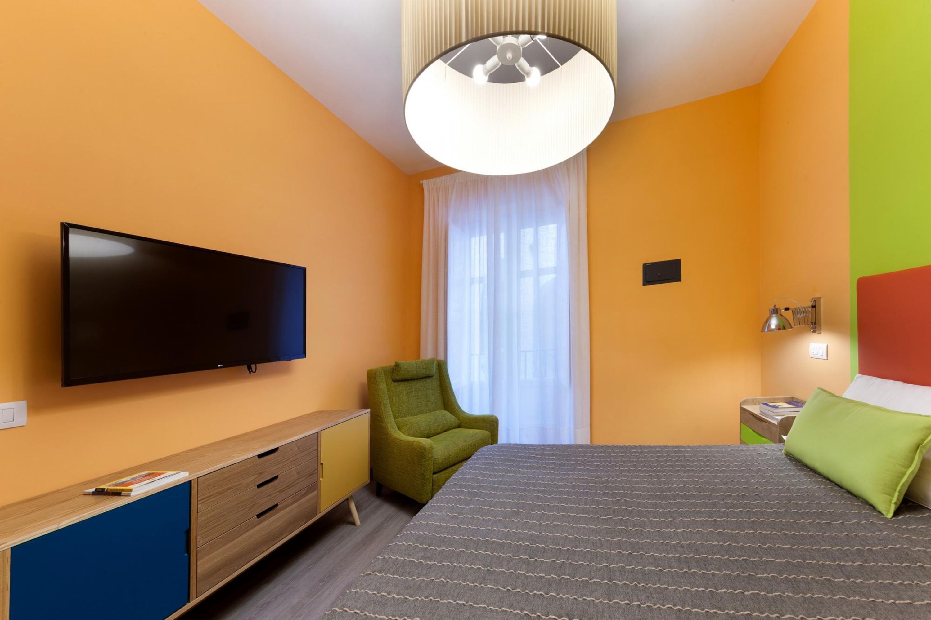A modern hotel room with a flat-screen TV, a green armchair, a bed with a patterned bedspread, a bedside table with a lamp, and a window with sheer curtains.