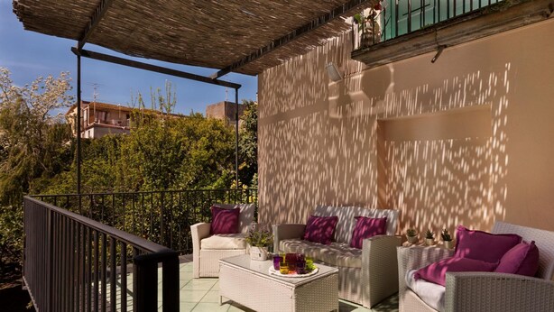 Un espacio al aire libre cubierto con mobiliario de mimbre y cojines, una mesa de centro con bebidas y vistas a zonas verdes y edificios.
