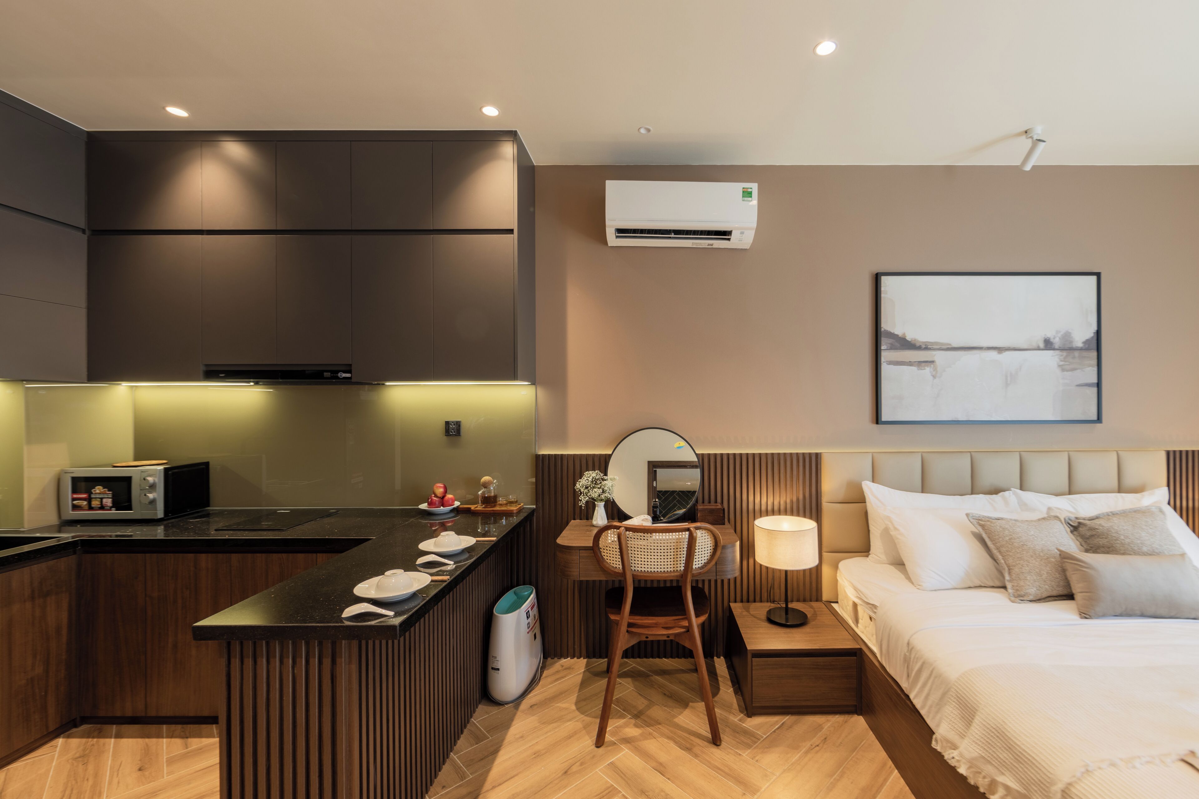 Quarto de hotel moderno com cozinha compacta, cama com roupa de cama branca, mesa de cabeceira com abajur e ar condicionado fixado na parede.