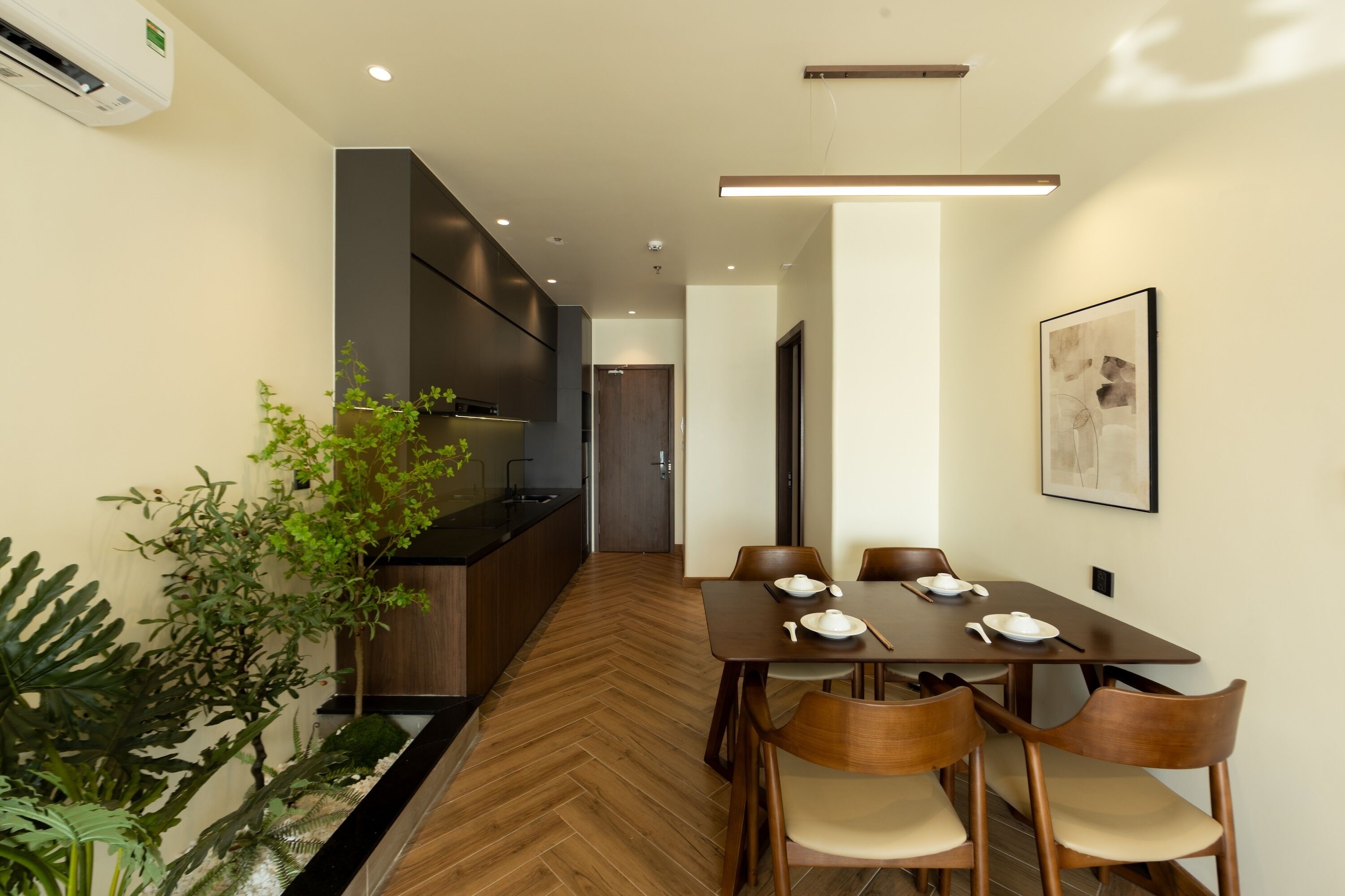 A modern dining area with a wooden herringbone floor, a long table with six chairs, and a kitchen area with a built-in stove and cabinets.