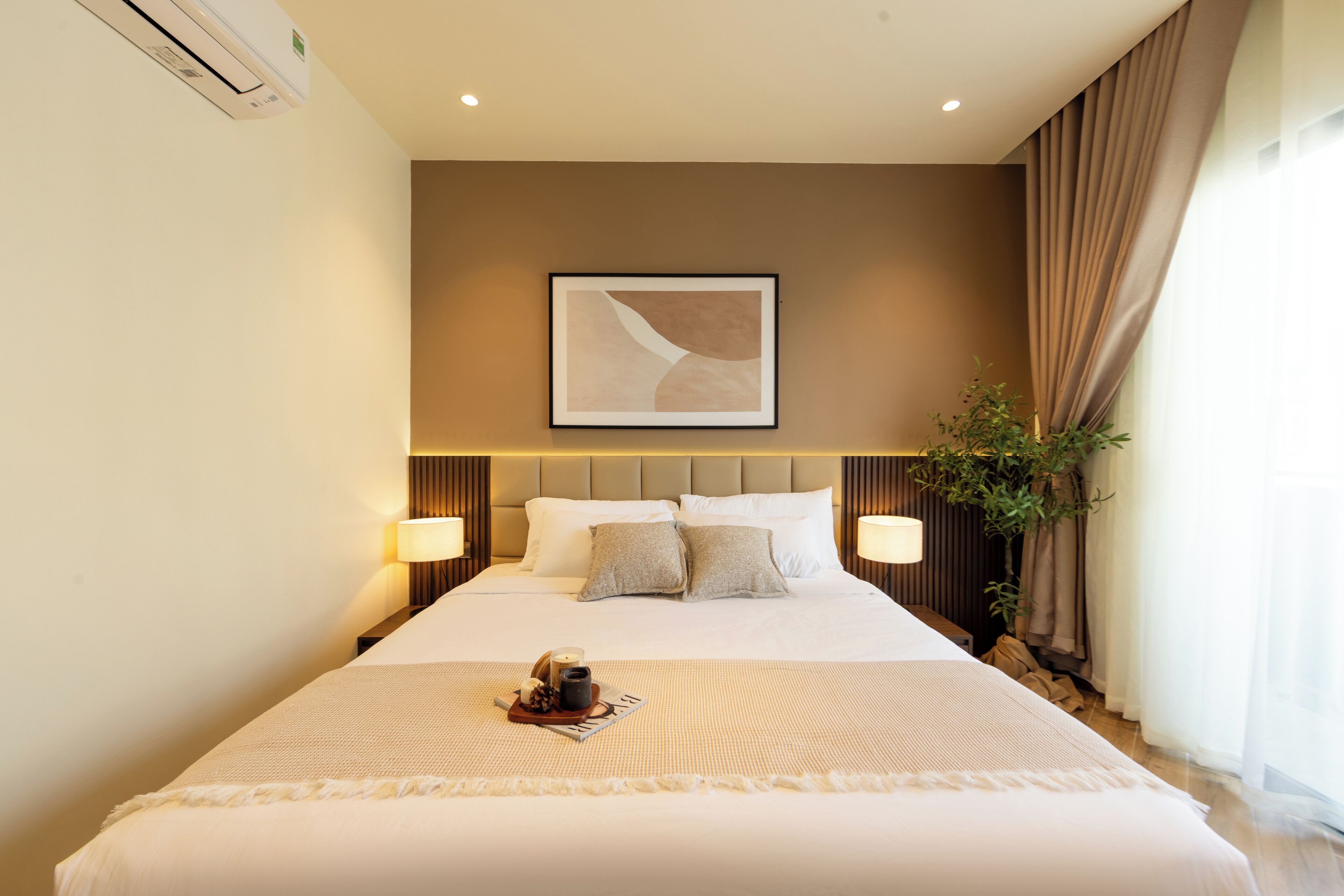 A hotel room with a large bed, two bedside lamps, a framed artwork above the bed, and a potted plant by the window.