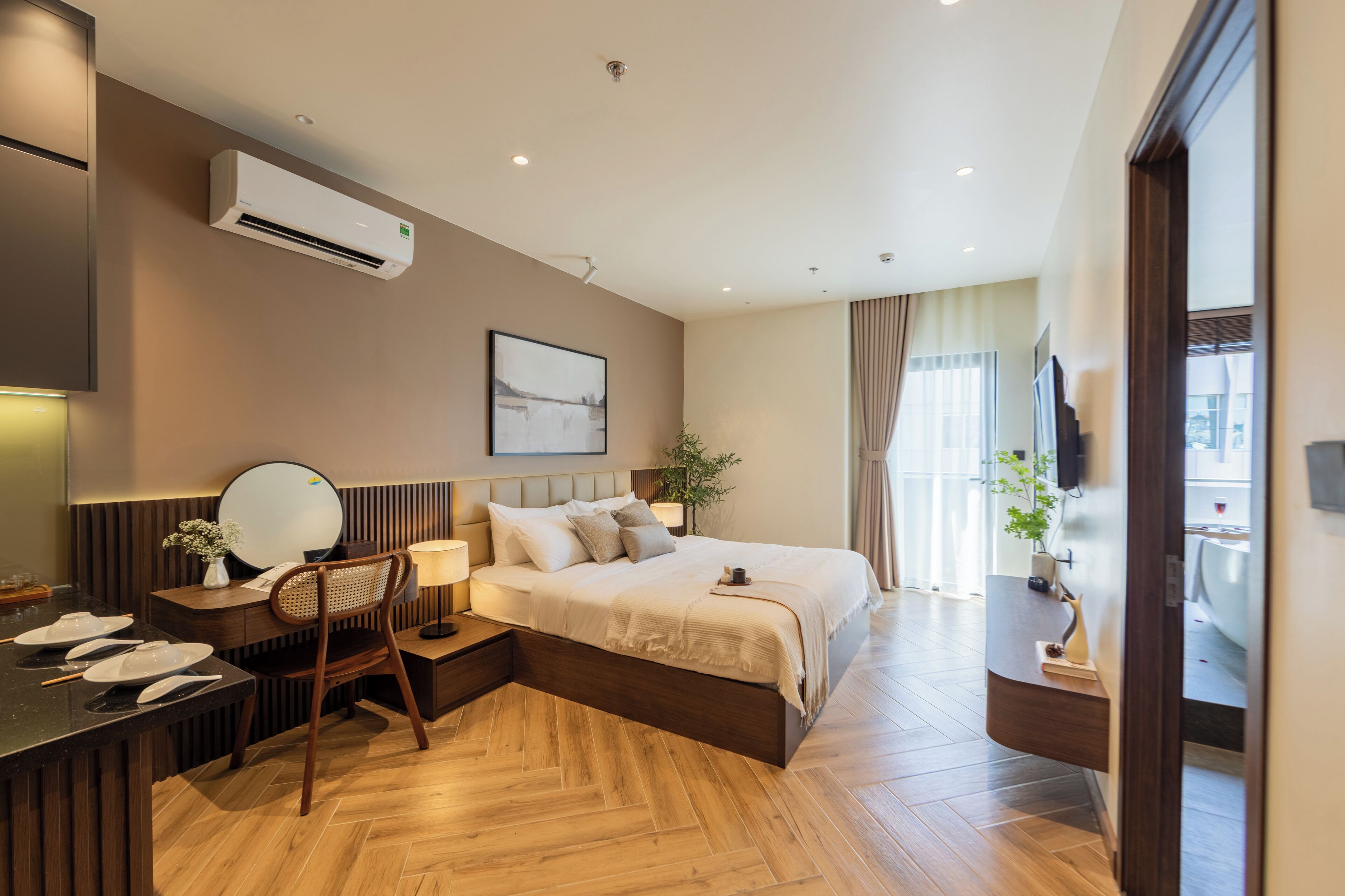 Una habitación de hotel moderna con piso de madera, una cama con ropa blanca, un escritorio con silla, una mesita de noche con lámpara y un aire acondicionado instalado en la pared.