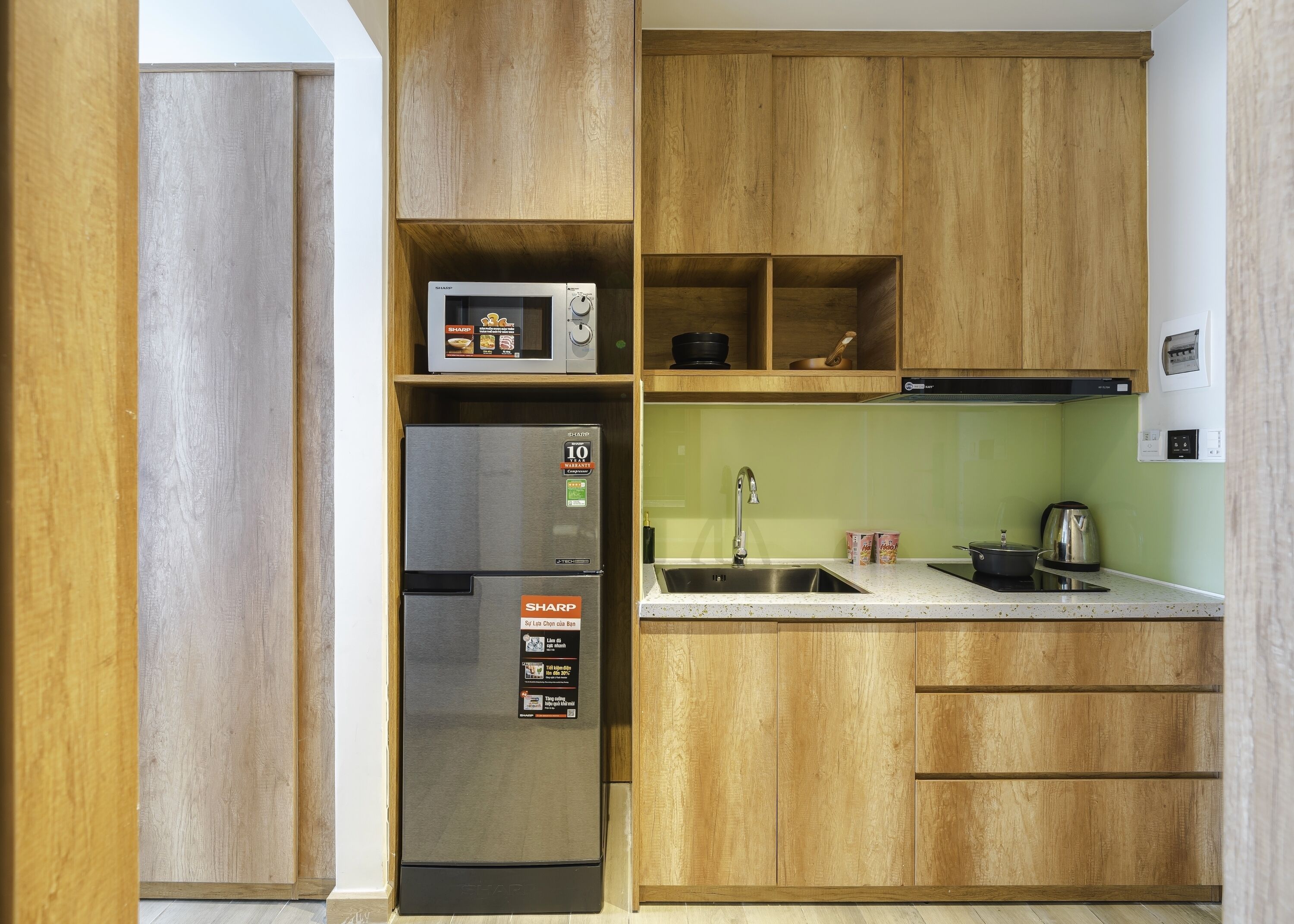 A compact kitchen with wooden cabinets, a stainless steel refrigerator, a microwave, and a sink with a kettle.