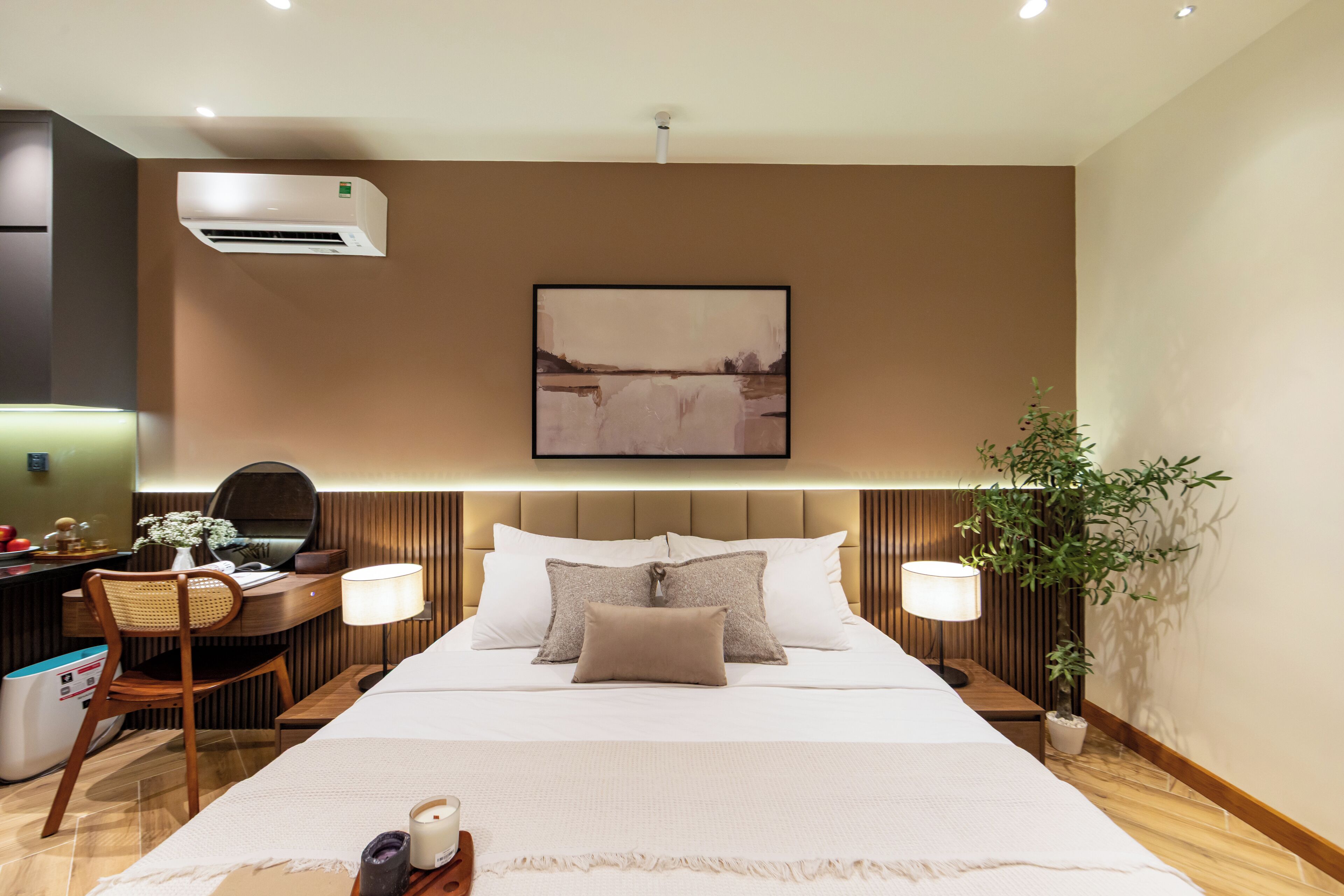 A modern hotel room with a large bed, bedside lamps, a desk with a chair, a potted plant, and an air conditioning unit.