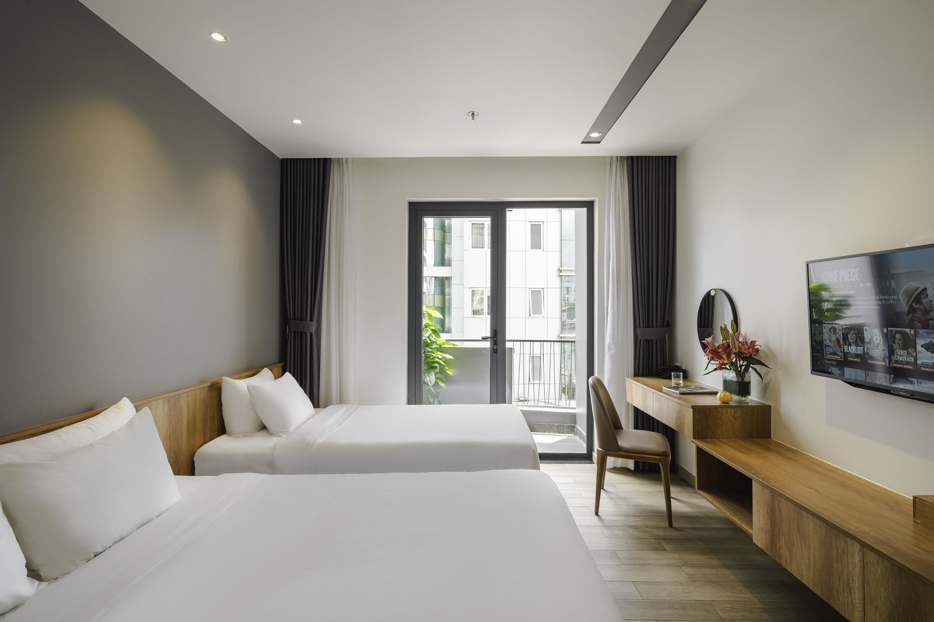A hotel room with two beds, a desk, a chair, a TV, and a balcony with a view of buildings.