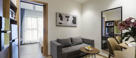 A modern living room with a grey sofa, a small round table with a bowl, a wooden dining table with chairs, a large mirror, and a painting on the wall.