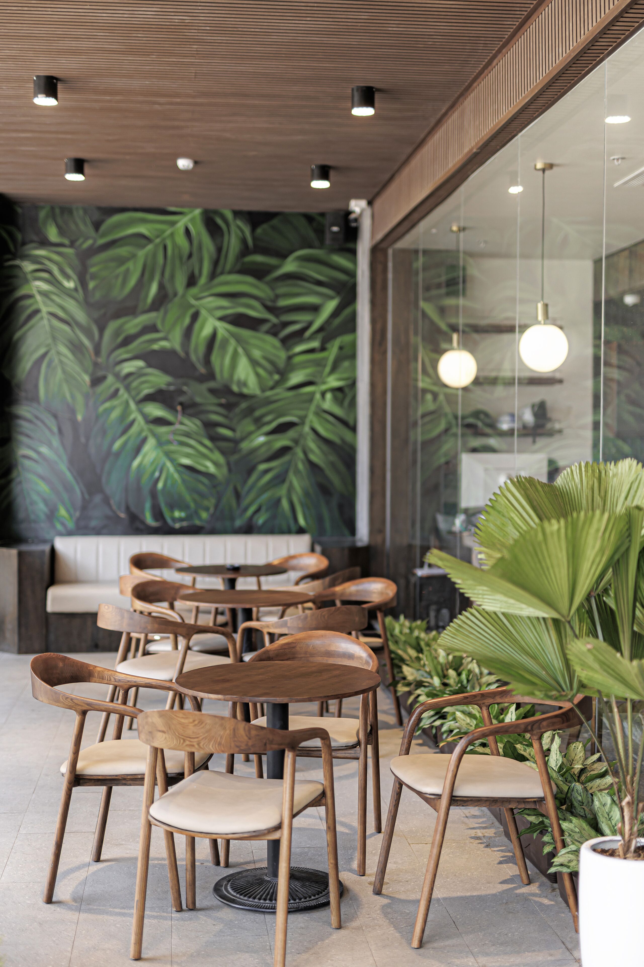 Een restaurantinterieur met houten stoelen, een muurschildering van groene bladeren en hangende pendellampen.