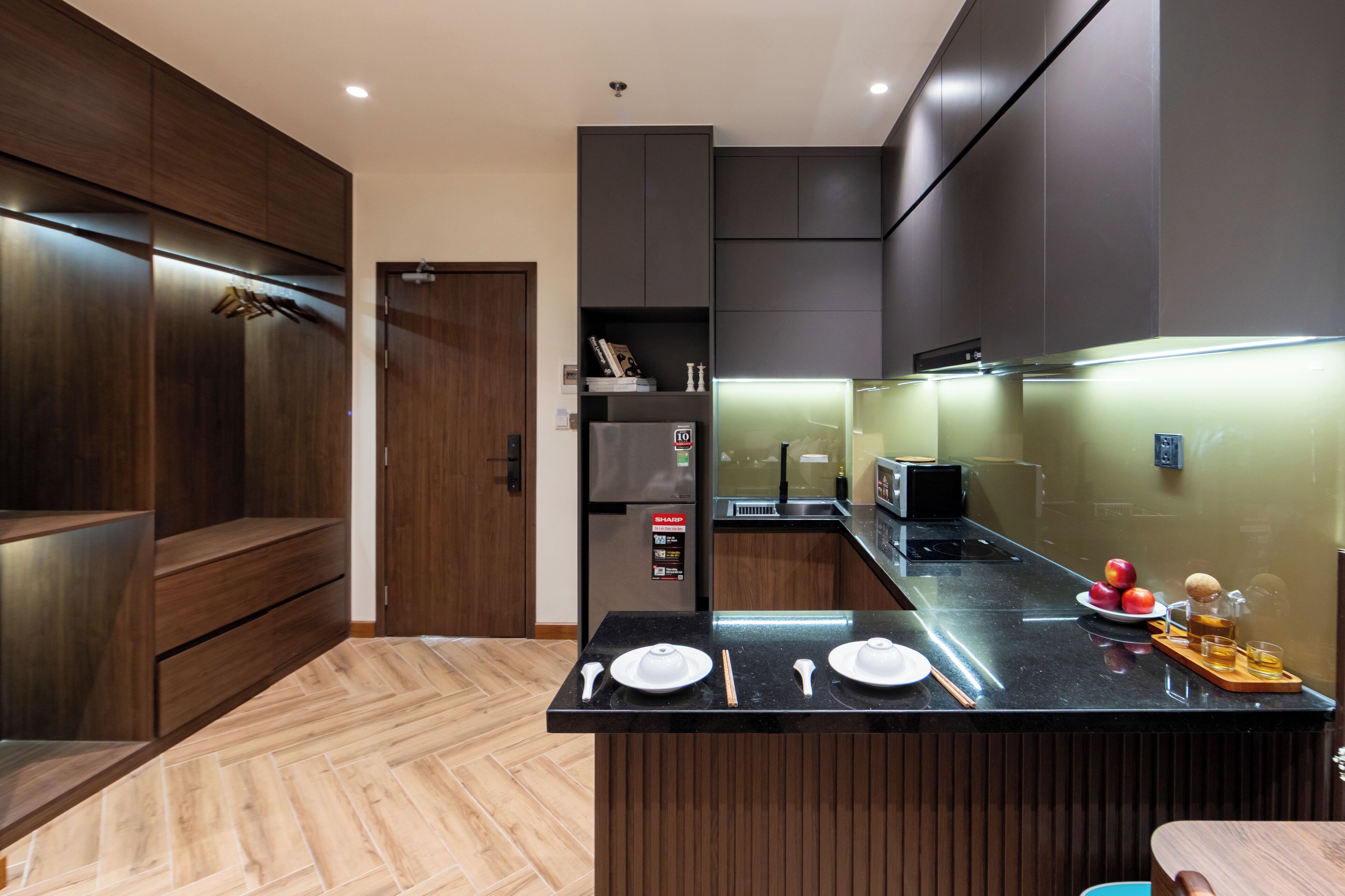 A modern kitchen with dark cabinetry, a wooden herringbone floor, and a central island with dining utensils and fruits.