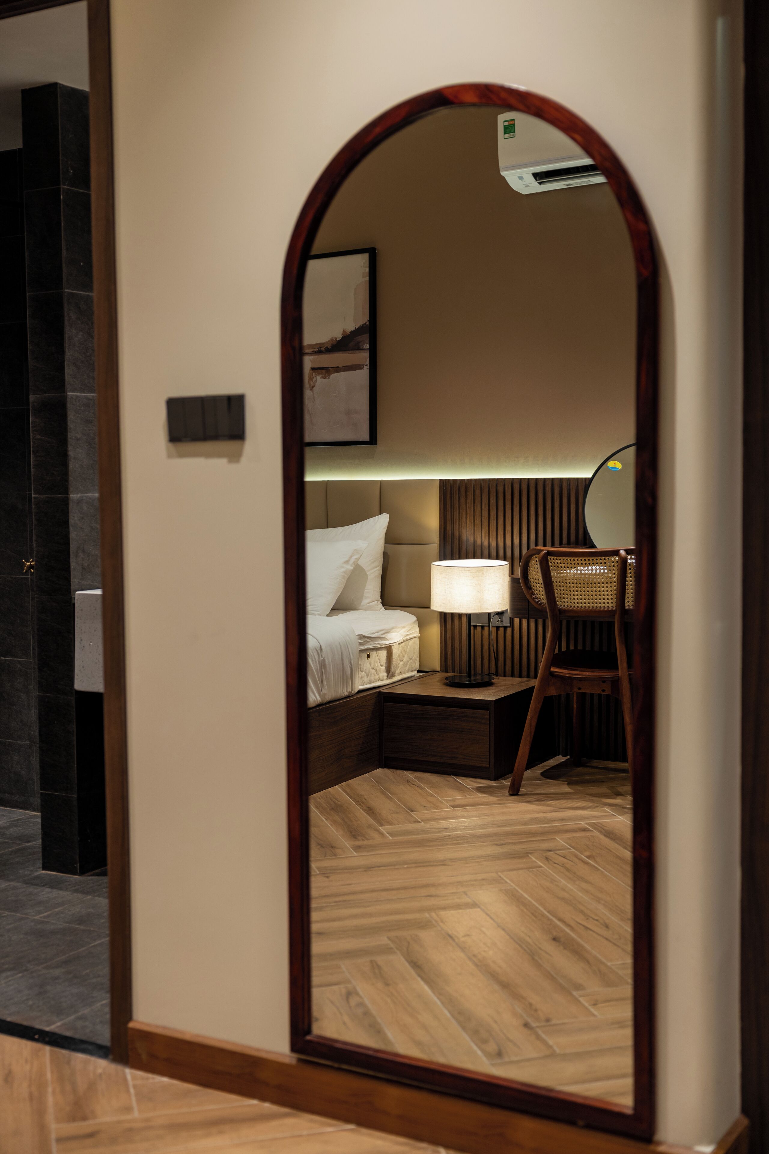 A hotel room with a wooden floor, a bed with white linens, a nightstand with a lamp, and a chair.
