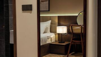 A hotel room with a wooden floor, a bed with white linens, a nightstand with a lamp, and a chair.