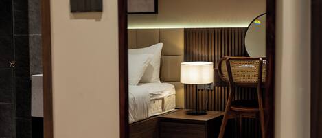 Habitación de hotel con piso de madera, cama con ropa blanca, mesita de noche con lámpara y una silla.