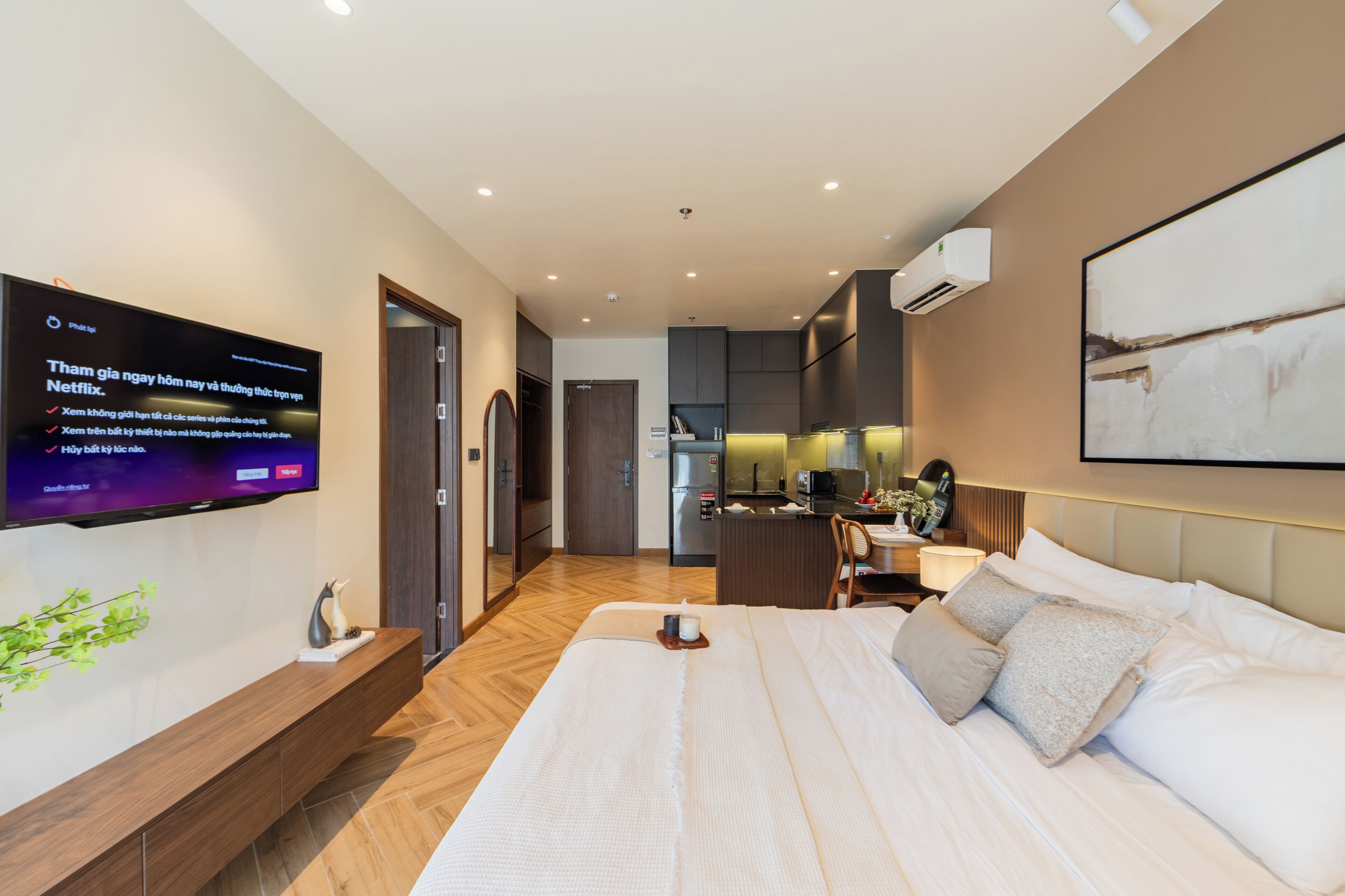 Moderni hotellihuone, jossa on suuri sänky, seinälle asennettu taulutelevisio, puinen konsolipöytä ja pieni keittotila.