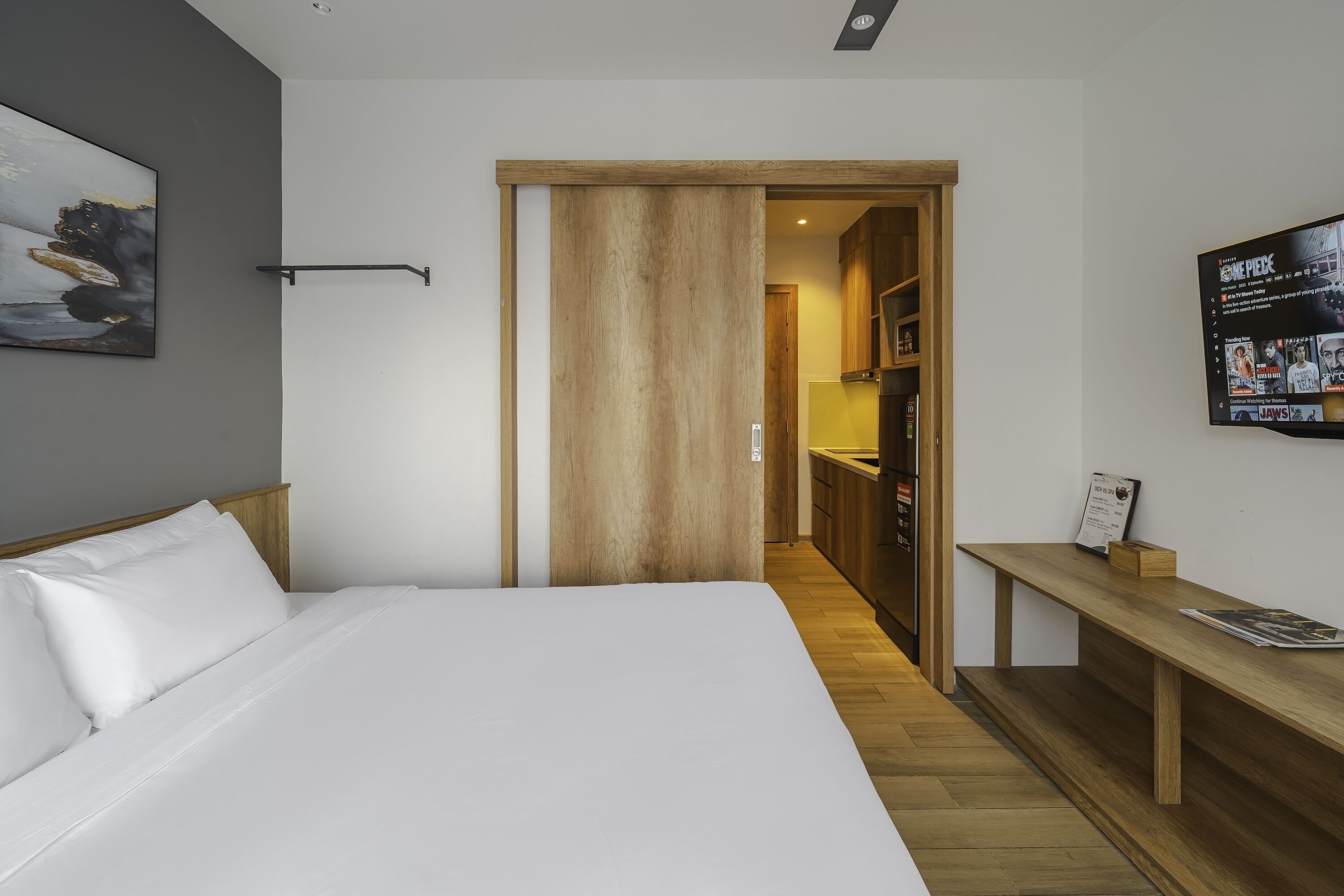 Una habitación de hotel moderna con una cama grande, una puerta de madera que da a una kitchenette y un televisor montado en la pared.