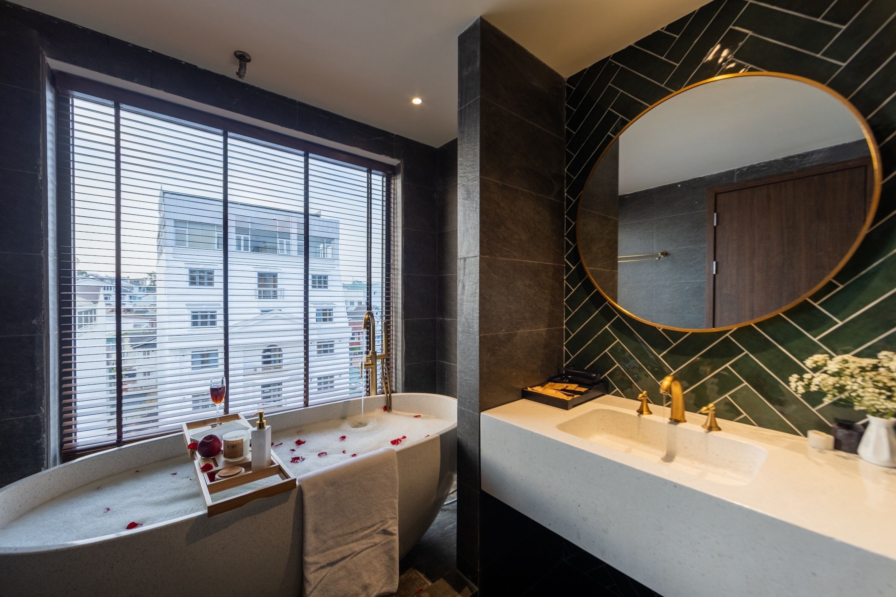 A modern bathroom with a freestanding bathtub, a large mirror, and a window with blinds.