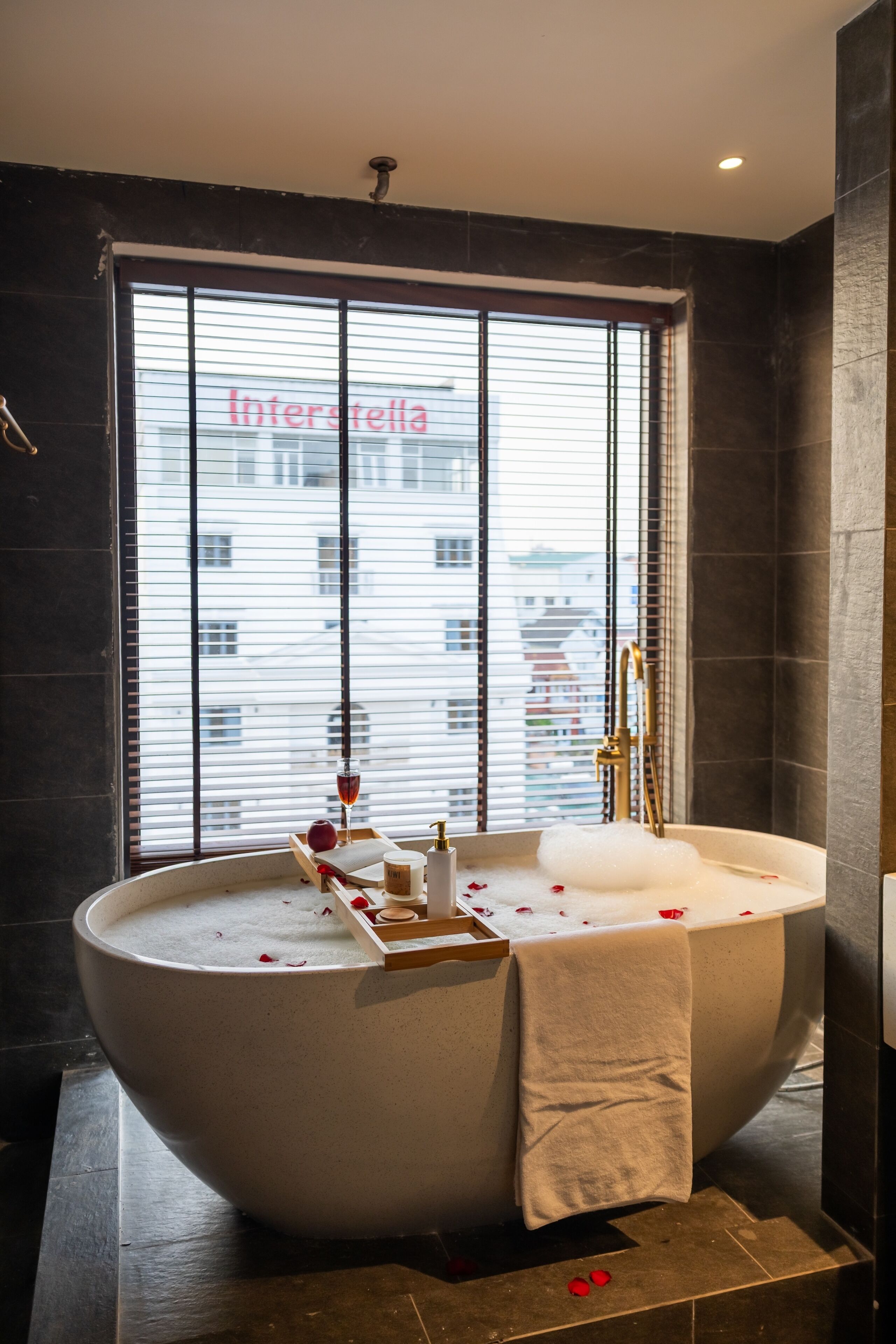 Une salle de bain moderne avec une baignoire sur pieds, une fenêtre avec des stores et une vue sur un immeuble.